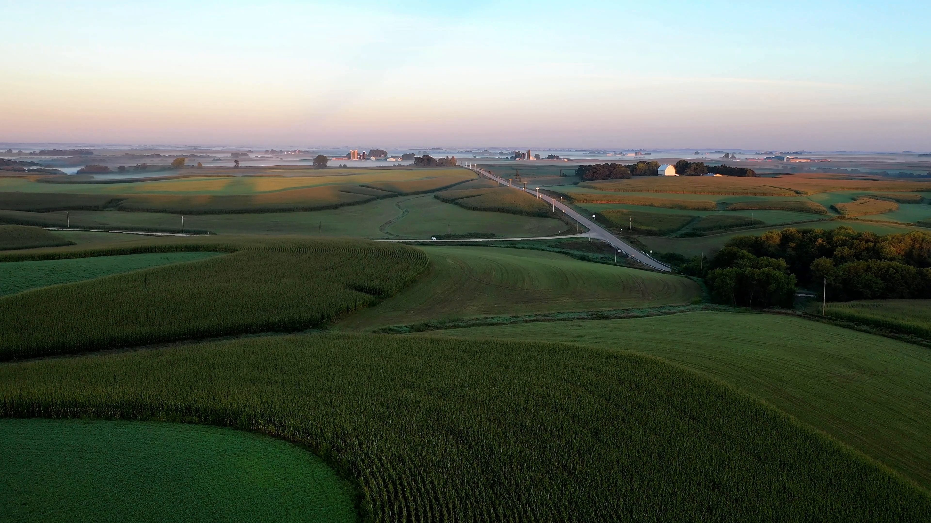 Vista aérea de lomas cultivadas al amanecer, con franjas sembradas en curvas de nivel, un camino rural, un granero blanco y silos a lo lejos, y neblina baja en el horizonte.