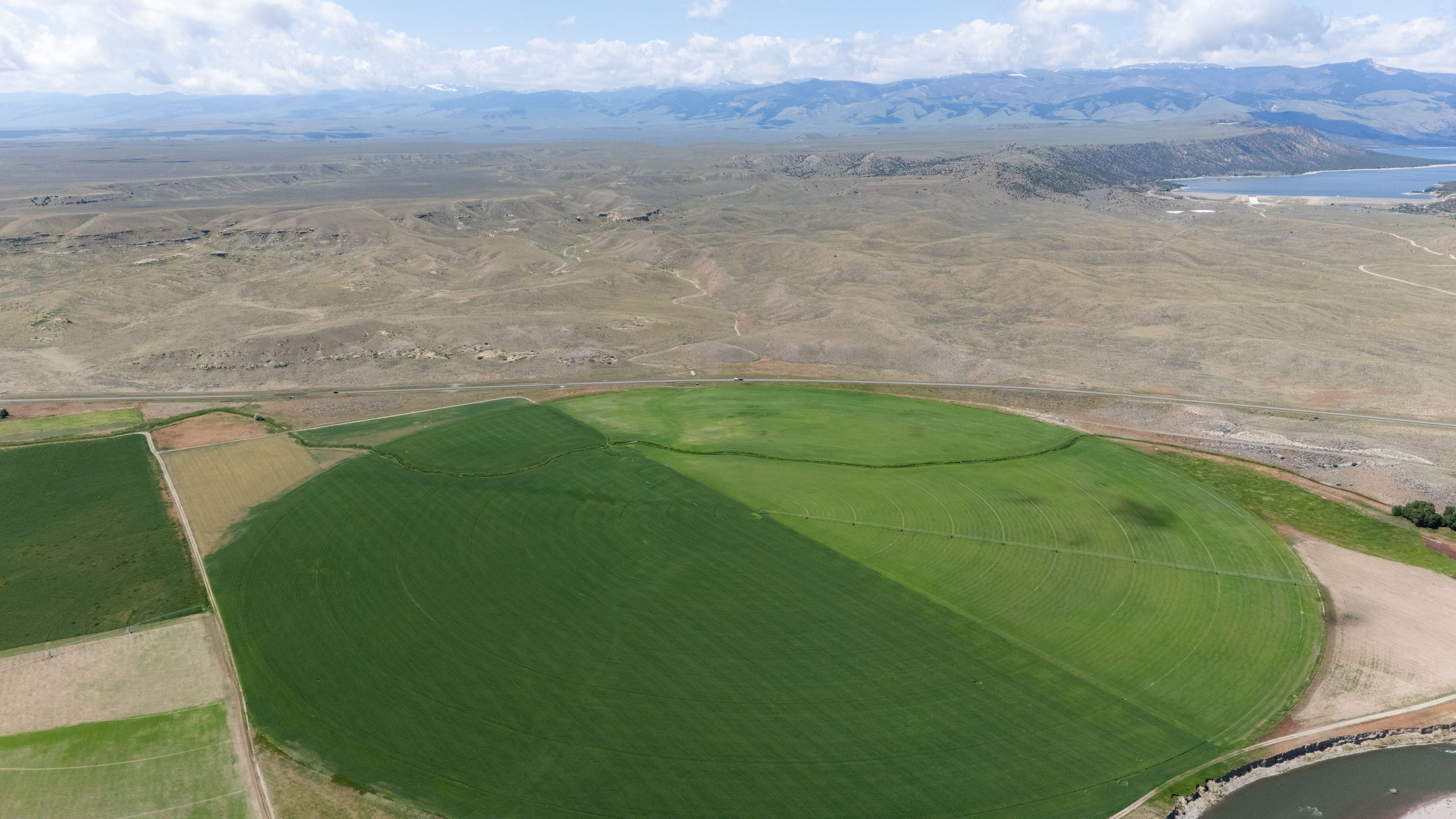 Vista aérea de un gran círculo verde regado por pivote central, con parcelas más pequeñas al lado, lomas secas, un embalse y montañas al fondo, bajo nubes.