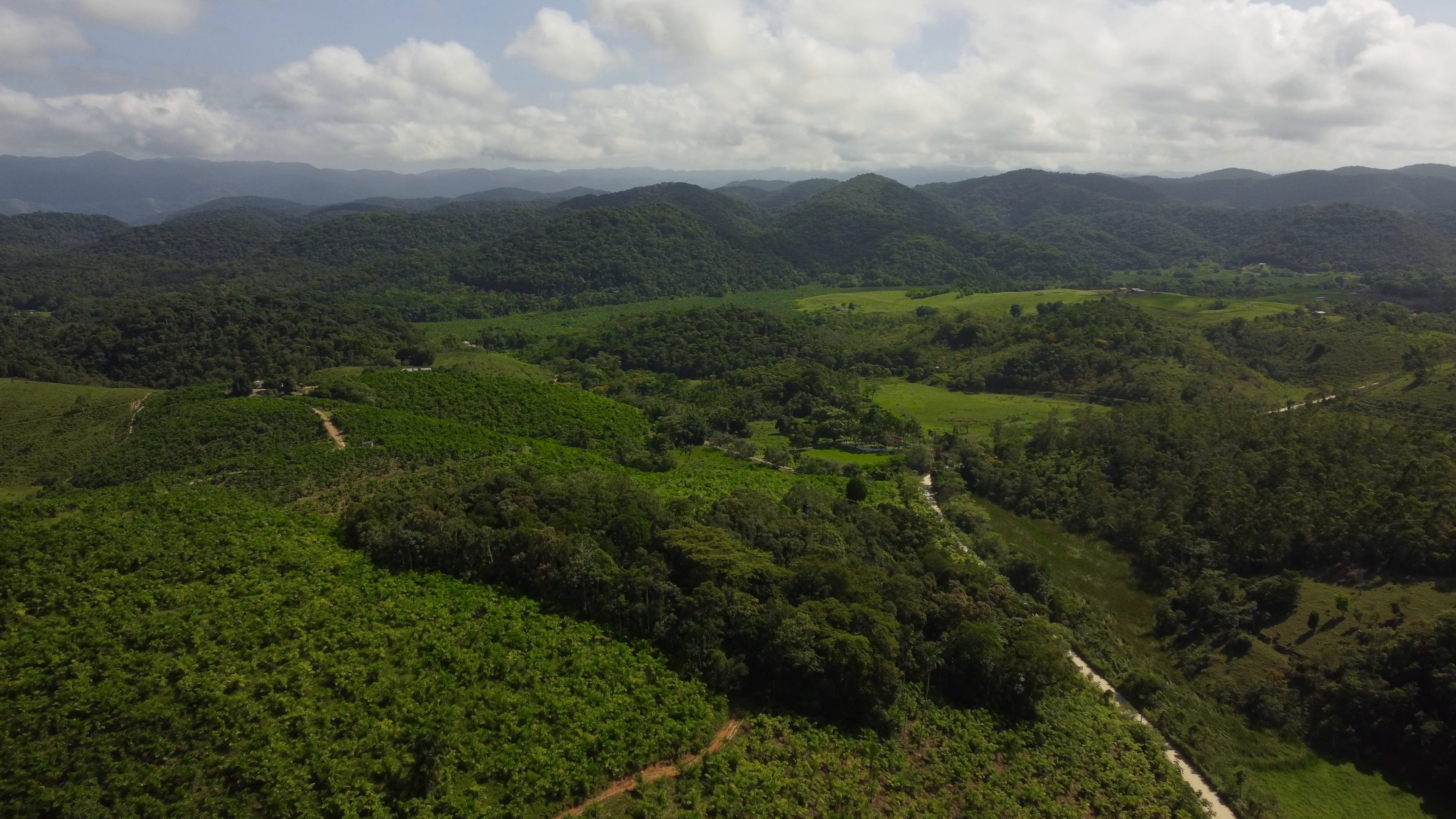 Vista aérea de morros no Brasil, com lavouras ao lado de manchas de mata, estradas de terra e pastos, e serras ao fundo sob nuvens.